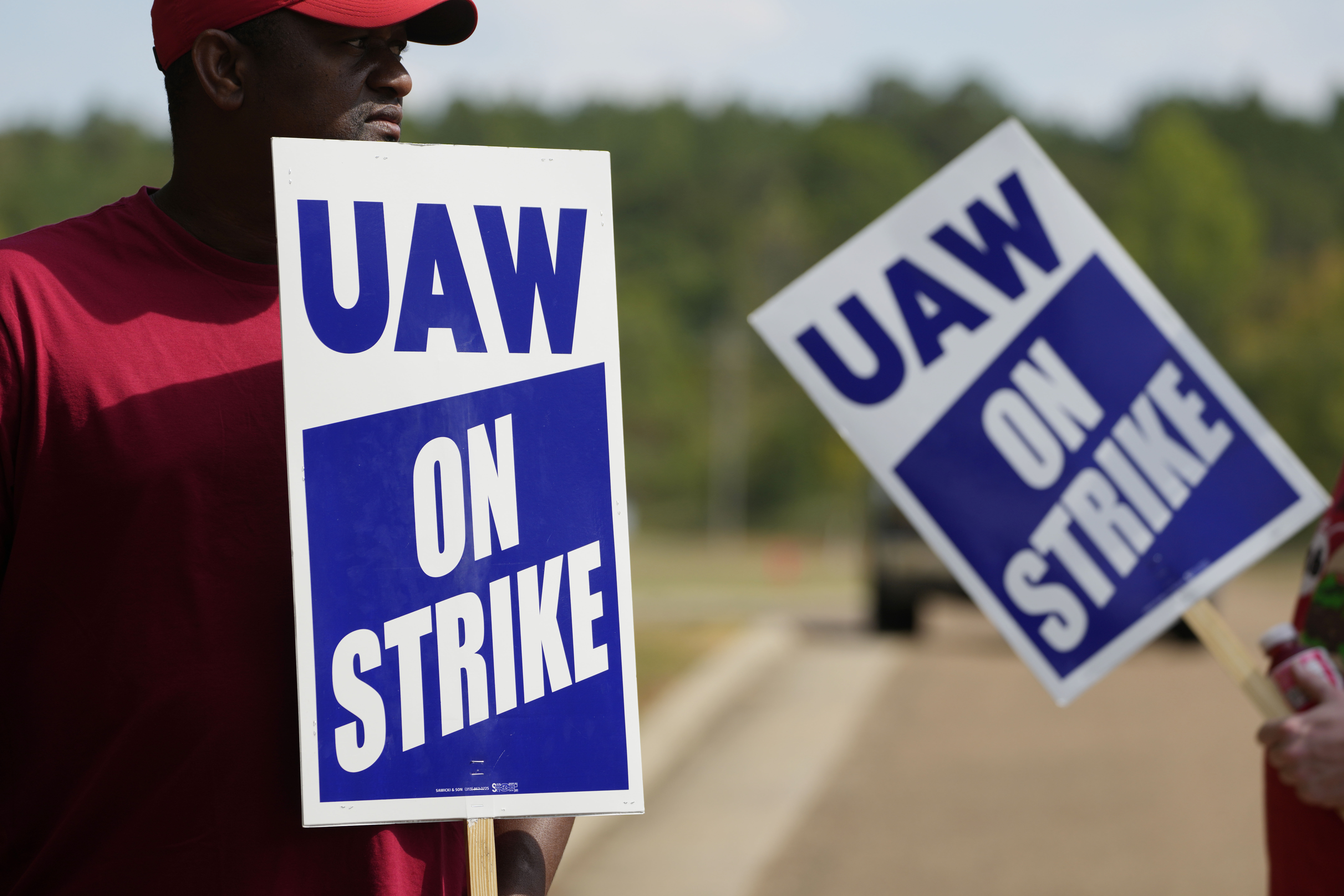 Image shows a man in a UAW shirt holding a large sign that says "UAW on Strike."