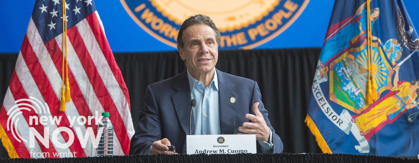 Governor sits at a press conference between the American and New York State flags.
